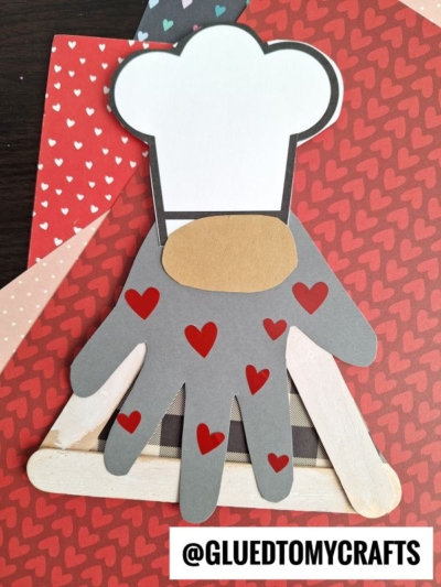 A paper craft of a baker gnome chef made from a gray handprint cutout with red hearts, a white chef hat, and a brown nose, placed on patterned heart paper and wooden popsicle sticks. Text reads @GLUEDTOMYCRAFTS.