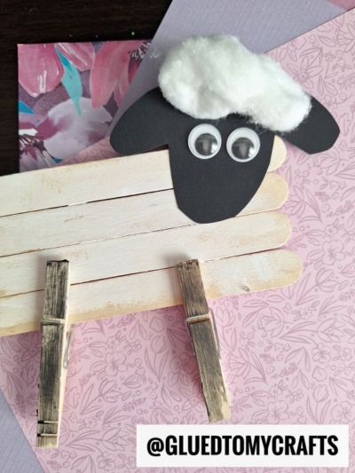 A popsicle stick sheep craft made from craft sticks and clothespins, with a black paper face, googly eyes, and cotton wool, placed on floral-patterned papers. @GLUEDTOMYCRAFTS is written at the bottom.