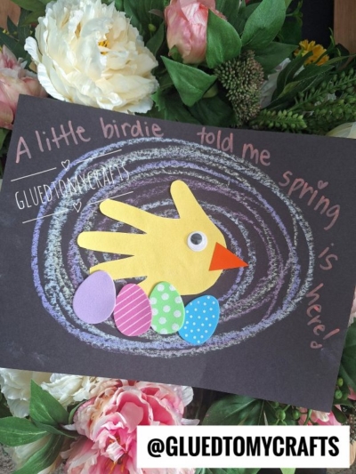 A handprint paper craft of a yellow bird with a red beak and orange wing, surrounded by four colorful eggs, sits on a black sheet with pink text. It looks like a cheerful chalk and paper bird celebrating spring!.