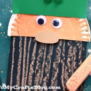A craft of a leprechaun face made from a paper plate, green paper hat, googly eyes, peach nose, and black paper with orange crayon lines for a chalk art leprechaun beard. A peach Crayola crayon lies nearby.