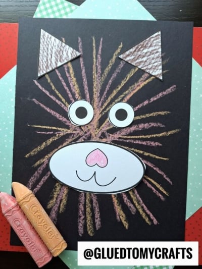 A vibrant chalk art cat face project with chalk lines for fur, googly eyes, a drawn nose and mouth on white paper, and textured triangle ears, displayed on layered colored paper. Two crayons and @GLUEDTOMYCRAFTS are visible nearby.