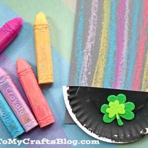 Four colorful sidewalk chalk sticks and a chalk art rainbow drawing on black paper. Next to it is a semicircle made from a painted paper plate decorated with a green shamrock. The text GluedToMyCraftsBlog.com appears at the bottom.