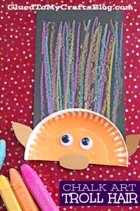 A colorful troll craft with chalk art troll hair on black paper, a half orange paper plate for the face, googly eyes, and tan paper ears. Bright chalk sticks and polka dot red fabric are also shown.