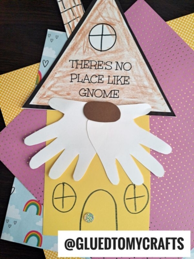 A handmade paper craft of a gnome house with a triangle roof displays the phrase "No Place Like Gnome," a brown door, yellow walls, round windows, and white handprint beards—all set against colorful backgrounds.