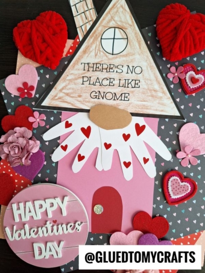 A handmade Valentine’s card features a gnome house with heart decorations, paper, felt, and yarn hearts on a dark background. Text reads “There’s no place like gnome” and “Happy Valentines Day.” No place like gnome for the holidays!.