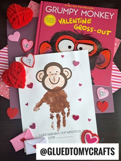 A children’s Valentine’s book titled “Grumpy Monkey Valentine Gross-Out,” a handprint monkey craft on white paper, felt and paper hearts, and a tag reading “@GLUEDTOMYCRAFTS” are arranged on a dark surface.