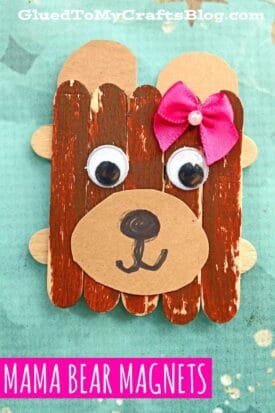 A popsicle stick mama bear magnet made of brown painted craft sticks and paper, with googly eyes, a pink bow on its ear, and a black nose and mouth drawn on the snout. Text reads, Mama Bear Magnets.