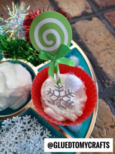 A red silicone cupcake liner holds white foam resembling moon dough slime with a snowflake decoration and a green lollipop stick, displayed on a tray with craft supplies. @GLUETOMYCRAFTS is written at the bottom.