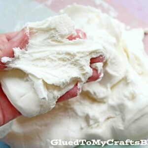 A hand kneads and stretches white, fluffy moon dough slime on a pastel surface. The texture appears soft and pliable. The words GluedToMyCraftsBlog.com are visible at the bottom of the image.