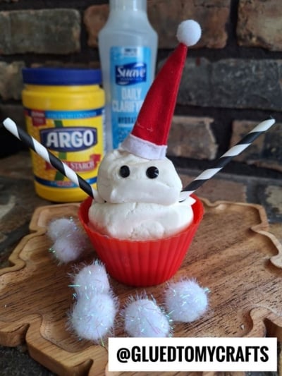 A snowman made of white slime dough with a red Santa hat, button eyes, and striped straws for arms sits in a red silicone cup. Fluffy pom-poms, cornstarch, and a bottle are in the background. @GLUEDTOMYCRAFTS appears in the corner.