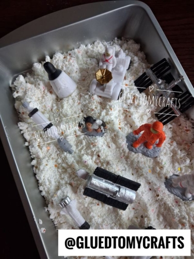 Toy astronauts, rockets, and vehicles are arranged on white sensory sand and shimmering moon slime in a baking dish. The scene mimics a moon or space exploration. An @GLUEDTOMYCRAFTS label appears at the bottom.