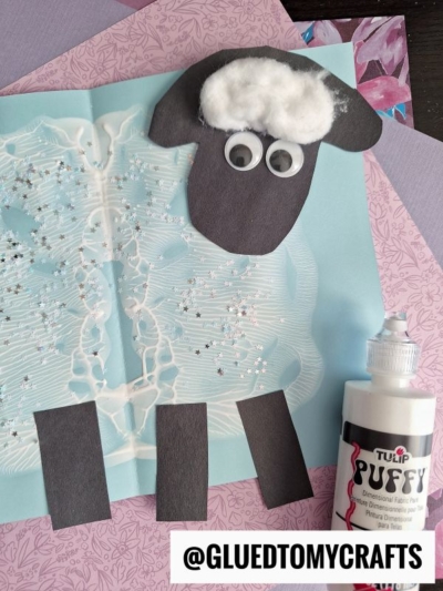 A sheep craft for kids made from black and white paper, cotton balls, and glitter on blue paper, with white puffy paint and craft supplies nearby. The Instagram handle @gluedtomycrafts is at the bottom.
