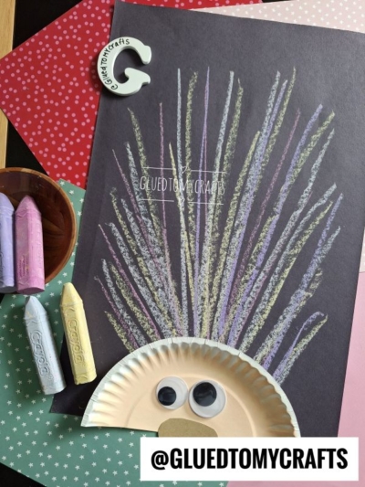A hedgehog craft made from a black paper background, colorful chalk art hair for spines, a paper plate for the face, and large googly eyes. Crayons, a glue stick, and patterned paper are beside the craft. Text: @GLUEDTOMYCRAFTS
