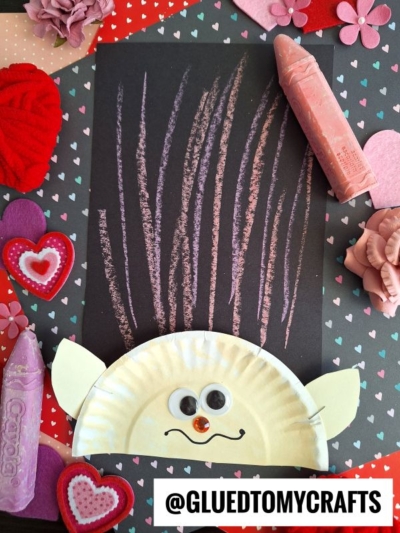 A paper plate troll craft with googly eyes, a red nose, yellow ears, and colorful chalk art hair sits on a black sheet with crayon marks, surrounded by heart decorations and large crayons on a heart-patterned surface. Text reads @GLUEDTOMYCRAFTS.