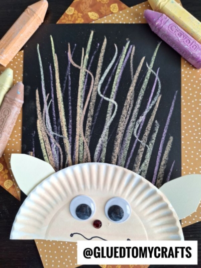 A kids hedgehog craft made with a paper plate, googly eyes, and chalk art hair for quills on black paper, surrounded by pastel crayons and orange patterned paper. @GLUEDTOMYCRAFTS is written at the bottom.