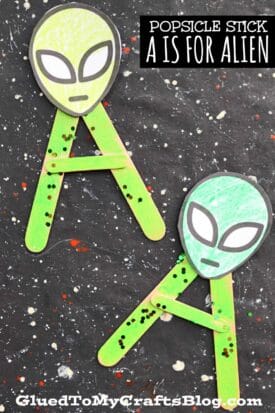 Two popsicle stick crafts shaped like the letter A, each topped with a cute popsicle stick alien face, rest on a black, star-speckled background. The text reads “POPSICLE STICK A IS FOR ALIEN.”.