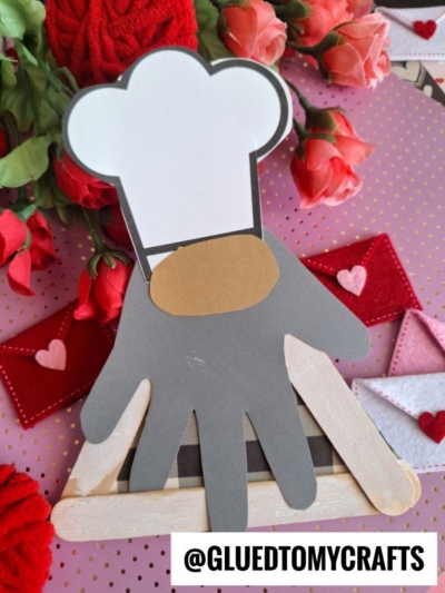 A handmade Valentine’s Day craft features a baker gnome with a white hat, gray hand-shaped body, and brown collar, framed by roses, pink hearts, and red and white felt envelopes. The handle @GLUEDTOMYCRAFTS is at the bottom.