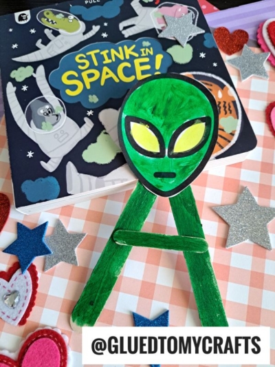 A children's book titled Stink in Space! is shown with a popsicle stick alien craft and green paper spaceship in front. Glittery stars and heart decorations surround them on a checkered surface. The text @GLUEDTOMYCRAFTS is at the bottom.
