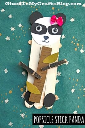 A popsicle stick panda bear craft with black paper ears, a pink bow, googly eyes, and a paper bamboo stick held by clothespins, displayed on a green star-patterned background. Text reads “Popsicle Stick Panda.”.