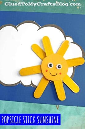 A cheerful popsicle stick sunshine craft with a smiling yellow sun, googly eyes, and a painted face sits on a white cloud cutout over a blue background. Text at the top reads GluedToMyCraftsBlog.com.