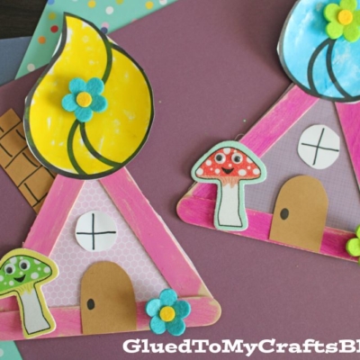 Two colorful, handmade craft stick troll houses, decorated with paper and felt flowers, sit on layered sheets of colored paper. Each fairy-inspired popsicle stick troll house features a mushroom decoration and a rounded door.