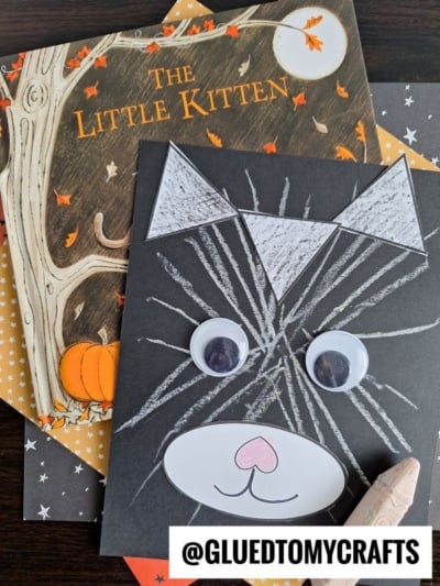 A handmade black chalk art cat with googly eyes and a pink nose sits atop the book “The Little Kitten,” surrounded by autumn-themed paper. A white crayon and the @GLUEDTOMYCRAFTS handle are also visible.