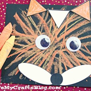 A chalk art fox craft made with orange and yellow crayon lines on black paper, large googly eyes, white paper shapes for the snout and ears, and three crayons beside the artwork. GluedToMyCraftsBlog.com appears at the bottom.