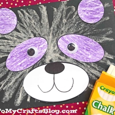A handmade chalk art panda face made from black paper, with white and purple chalk details. Crayola chalk, tape roller, and part of a pen are beside this fun panda craft for kids on a red polka-dot background. GluedToMyCraftsBlog.com is at the bottom.