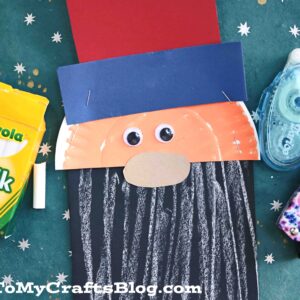 A Uncle Sam craft made with a paper plate, colored paper, googly eyes, and chalk art details is surrounded by chalk, tape, and a stapler on a green surface. GluedToMyCraftsBlog.com is written at the bottom.
