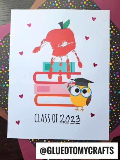 A card featuring stacked books, an owl in a graduation cap, a handprint apple and Class of 2023 text. Small hearts and colorful star paper surround the card, perfect for celebrating your handprint class of graduates. @GLUEDTOMYCRAFTS is at the bottom.