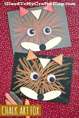 Two chalk art fox faces are drawn with orange chalk on black paper, featuring large googly eyes and white paper details. The red background has scattered yellow dots and visible chalk sticks. Text reads "Chalk Art Fox.
