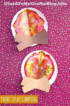 Two paper plate crafts resembling paint splat campfires, featuring vibrant paint-splattered “flames” and brown paper log strips, displayed on a pink polka-dot background. Text reads “PAINT SPLAT CAMPFIRE.”.