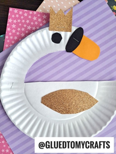 A paper plate swan craft with a gold glitter crown and wing, orange beak, and black oval detail is displayed on pastel patterned paper backgrounds. The handle @GLUEDTOMYCRAFTS is at the bottom.