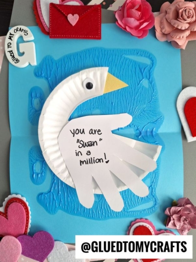 A handmade card features a swan made from a white cupcake liner, inspired by a paper plate swan craft, and a traced hand on blue paper, surrounded by hearts. The swan’s body reads: “You are ‘Swan’ in a million!”.