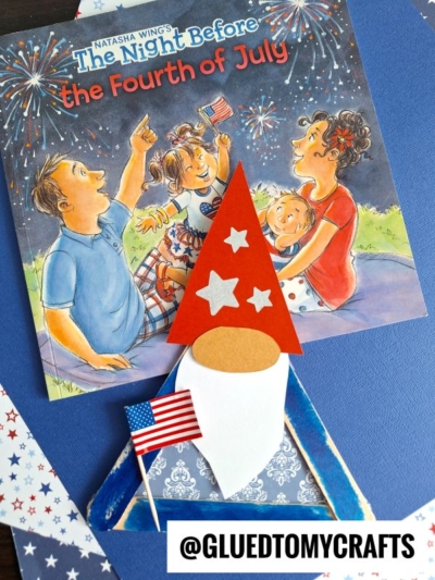 A children's book titled The Night Before the Fourth of July is displayed on a table with a popsicle stick patriotic gnome craft wearing a red hat and holding a small American flag in front. Patriotic star-patterned paper surrounds them.