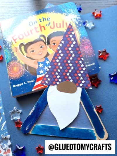 A popsicle stick patriotic gnome with a star-patterned hat, white beard, and blue triangle body sits on a table near a children’s book titled On the Fourth of July. Red, white, and blue star decorations are scattered around.