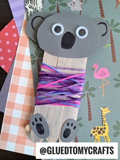A popsicle stick koala craft made with gray paper, googly eyes, and purple yarn sits on a background with animal illustrations. The image is labeled @GLUEDTOMYCRAFTS.