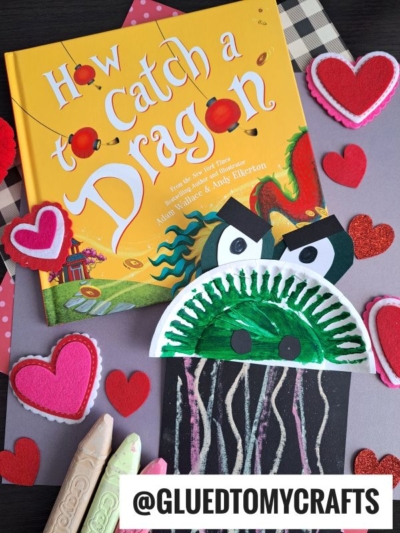 A children’s book titled How to Catch a Dragon is surrounded by felt and glitter heart decorations. Below the book is a dragon craft made from painted paper plates, with nearby large chalks ready for chalk art dragon creations.