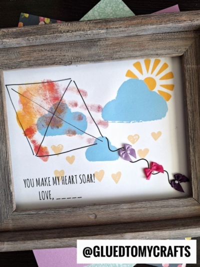 A framed craft features a handprint kite with finger-paint marks, blue clouds, yellow sun and hearts, plus a string with ribbon bows. Text reads: You make my heart soar! Love, ___. @GLUEDTOMYCRAFTS is at the bottom.