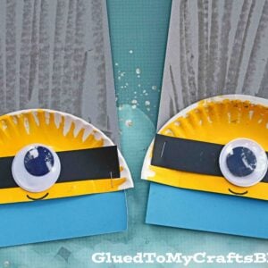 Two paper plate minion faces crafted from yellow plates, black strips for goggles, large googly eyes, and blue paper at the bottom, set on a teal background. The image features the text GluedToMyCraftsBlog.com.