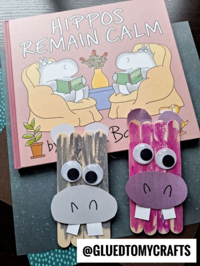 A children’s book titled Hippos Remain Calm is shown with two popsicle stick hippo crafts featuring googly eyes and paper accents. The hippos are brown and purple. @GLUEDTOMYCRAFTS is written at the bottom.