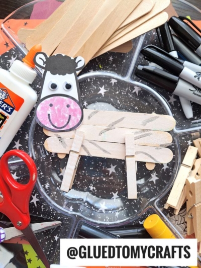 A cow craft made from popsicle sticks, paper, and googly eyes is surrounded by fun supplies like glue, markers, scissors, and clothespins on a star-patterned tray. A popsicle stick zebra could also be created. The handle @GLUEDTOMYCRAFTS is visible.