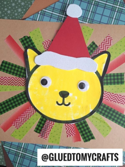 A handmade yellow cat face with a red Santa hat is centered on a brown background, surrounded by a washi tape lion mane made from green, red, and patterned tape strips. @GLUEDTOMYCRAFTS is written at the bottom.