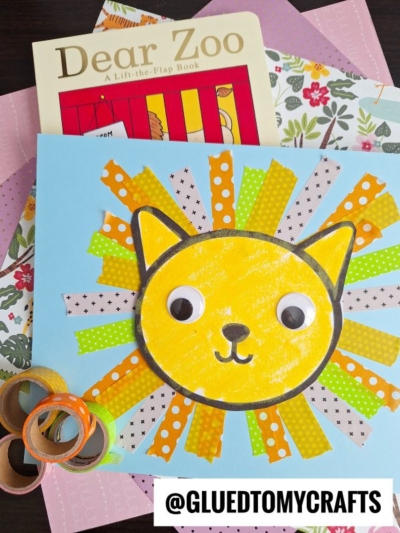 A children’s craft features a yellow cat face with a washi tape lion mane and googly eyes. Behind it are patterned papers, a Dear Zoo book, and rolls of washi tape. @GLUEDTOMYCRAFTS is written below.