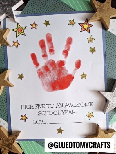 A red high five handprint on a paper says, High five to an awesome school year! Love, ____. Gold stars decorate the page, which is framed with blue paper. Stars also surround the paper on the table.