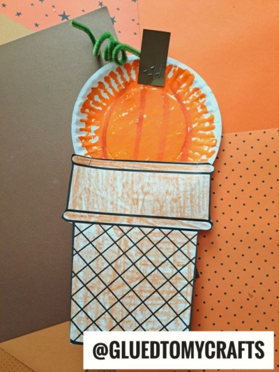 A paper craft torch with an orange flame and brown cone, held together by a green pipe cleaner and gold clip, lies on layered orange and brown paper—perfect inspiration for your next paper plate pumpkin ice cream project. @GLUEDTOMYCRAFTS is written at the bottom.