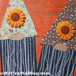 Two handmade chalk art fall gnome crafts feature patterned paper hats, felt sunflowers, and chalk-drawn beards on black paper. Displayed on an orange textured surface with colorful chalk pieces, they brighten up any autumn décor.