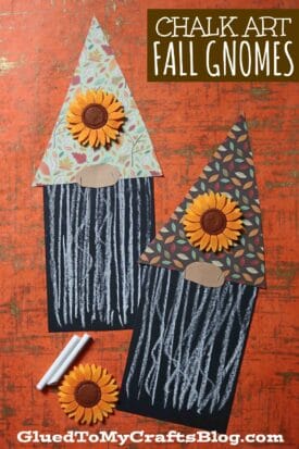 Two handmade chalk art fall gnome crafts feature patterned paper hats, felt sunflowers, and chalk-drawn beards on black paper. Displayed on an orange textured surface with colorful chalk pieces, they brighten up any autumn décor.