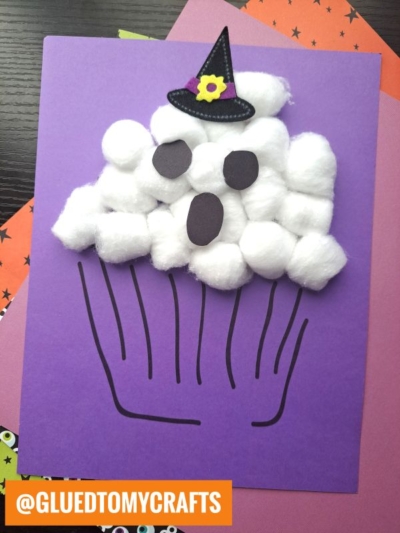 A cotton ball ghost cupcake outlined on purple paper, decorated with fluffy cotton balls as icing, black paper eyes and mouth, and a small witch hat perched on top. An orange label reads @GLUEDTOMYCRAFTS.