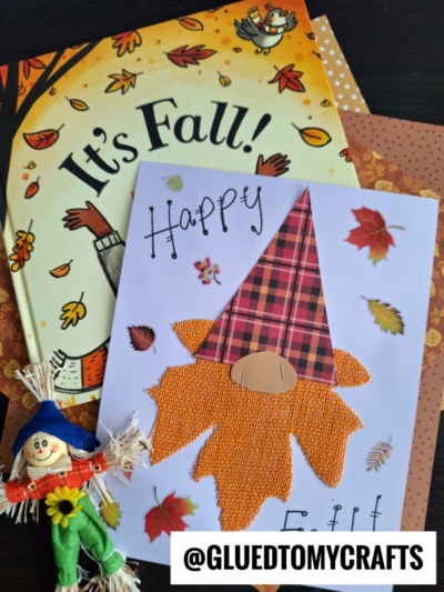 A handmade fall card featuring a plaid and burlap leaf gnome craft is displayed on autumn-themed papers and an “It’s Fall!” book. A small scarecrow figurine and scattered leaf stickers sit beside the card. Text: @GLUEDTOMYCRAFTS.